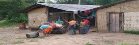 A group of African boys wearing colourful t-shirts, siphoning petrol from metal drums next to two wooden huts.